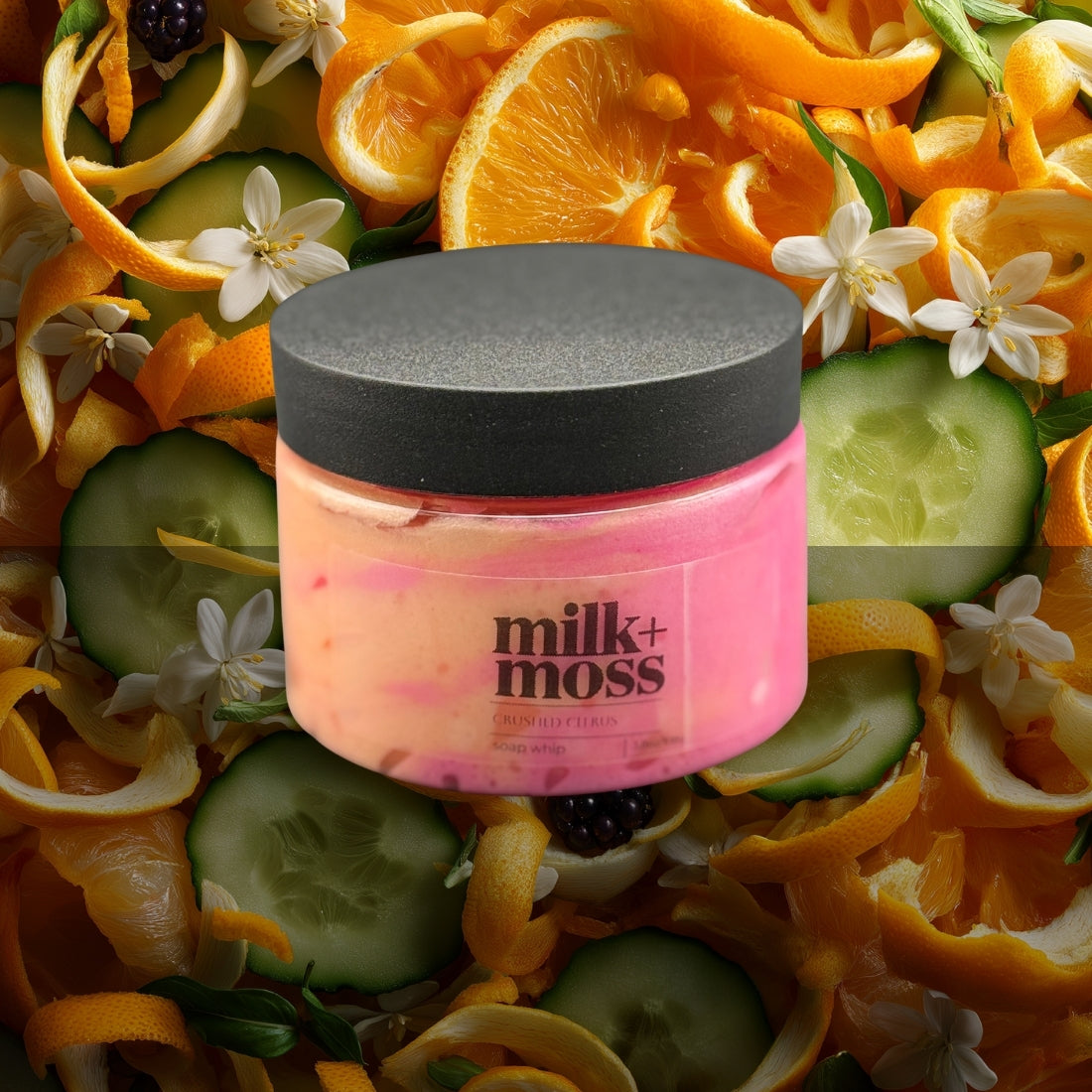 Pink jar with black lid labeled 'milk+moss' surrounded by orange slices, cucumbers, and flowers.