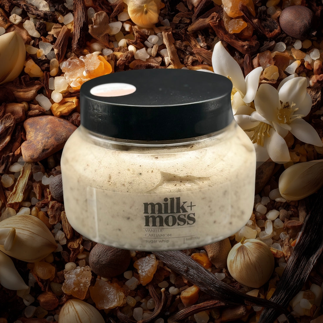 Jar of 'milk+moss' body scrub surrounded by natural elements like flowers and stones.