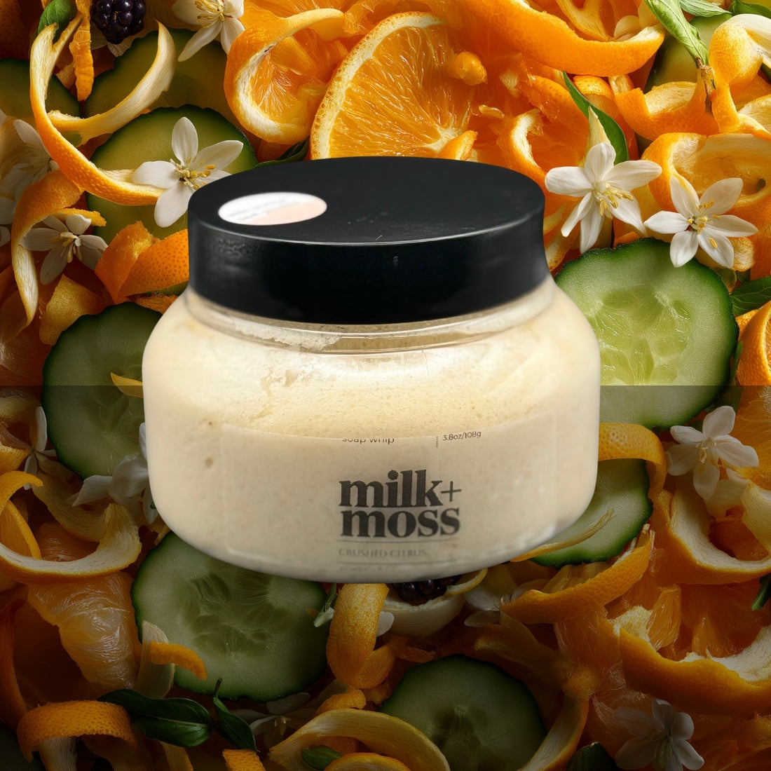 Jar of 'milk+moss' scrub surrounded by orange peels, cucumbers, and flowers.